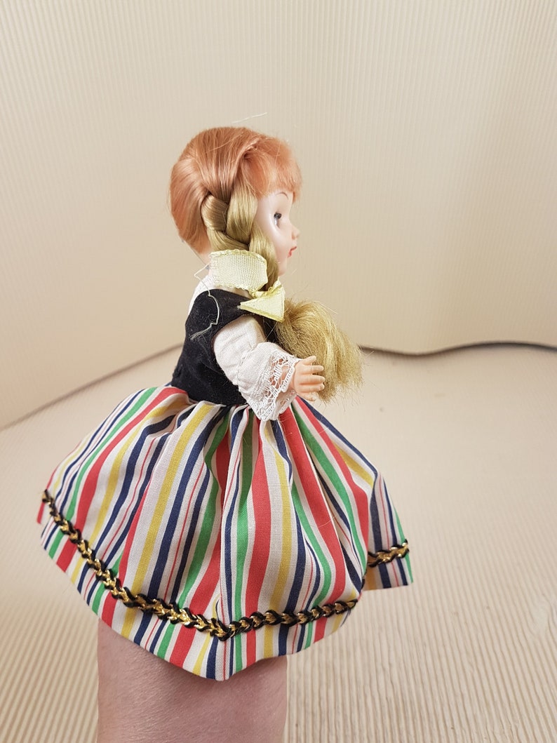 May include: A small doll with blonde hair in a braid, wearing a black bodice and a colorful striped skirt with gold trim.