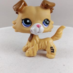 Puede incluir: Un perro de juguete de plástico marrón y beige con ojos azules y una nariz rosa. El perro de juguete tiene orejas marrones y una cola esponjosa. Las letras "LPS" están impresas en la pata del perro. Es un objeto de colección.
