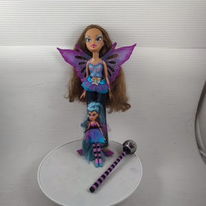 May include: Two Bratz dolls, one large and one small, both with fairy wings. The larger doll has brown hair and wears a blue top, a purple skirt, and butterfly wings. The smaller doll has blue hair and a similar outfit. A black and purple wand is also present.