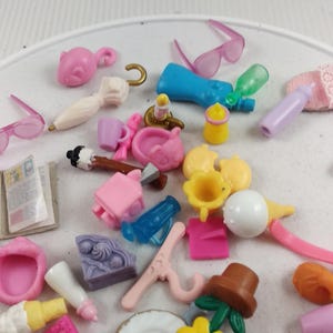 Puede incluir: Una colecci&oacute;n de accesorios de casa de mu&ntilde;ecas en miniatura en varios colores. Incluye peque&ntilde;as gafas de sol, botellas, un plato con dise&ntilde;o floral, una tetera y otros juguetes peque&ntilde;os. Los art&iacute;culos est&aacute;n dispersos sobre una superficie blanca.