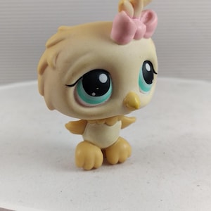May include: A small, tan-colored bird figurine with large, teal-colored eyes and a pink bow on its head. The toy bird has a yellow beak and feet, and is standing on a white surface. The figurine is a collectible toy.