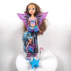 Puede incluir: Dos muñecas estilo Bratz, una grande y otra pequeña, ambas con alas de hada. La muñeca más grande tiene cabello castaño, un top rosa y azul y jeans. La muñeca más pequeña tiene cabello azul y un atuendo morado y azul. Accesorios: estrella azul y botella.