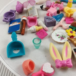 Puede incluir: Una colecci&oacute;n de accesorios en miniatura para casas de mu&ntilde;ecas en varios colores, que incluyen una cuchara azul con un recorte en forma de coraz&oacute;n, un cepillo para el pelo rosa y orejas de conejo amarillas con un lazo rosa. Otros art&iacute;culos incluyen un plato con un dise&ntilde;o floral y un libro en miniatura.