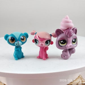 May include: Three colorful, small plastic animal figurines. A blue bear-like figure, a pink monkey-like figure, and a purple cat-like figure with a pink swirl on its head. Each has large eyes and is in a sitting position.