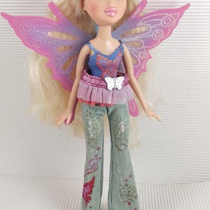 May include: A Bratz doll with long blonde hair and large eyes, wearing a blue top, pink butterfly wings, and green jeans with butterfly and floral designs. The doll has blue shoes.