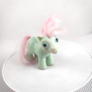 Puede incluir: Un juguete de poni vintage de color verde claro con una melena y cola rosas. El poni tiene ojos azules y un hocico blanco. El juguete está sobre una superficie blanca.
