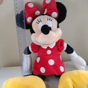 May include: A plush red and white polka dot Minnie Mouse toy with yellow shoes and a black bow. The toy is sitting on a white surface.