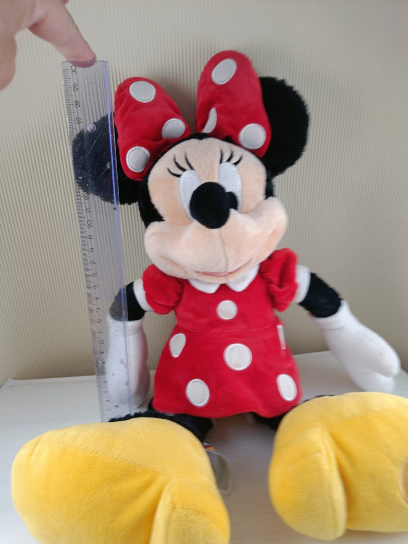 May include: A plush red and white polka dot Minnie Mouse toy with yellow shoes and a black bow. The toy is sitting on a white surface.