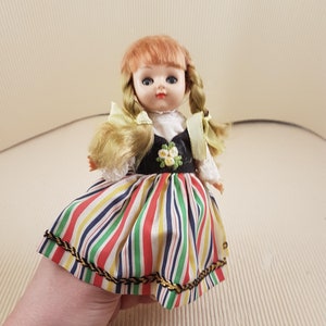 May include: A vintage doll with blonde hair in pigtails, wearing a white blouse, a black bodice with embroidered flowers, and a colorful striped skirt with gold trim.
