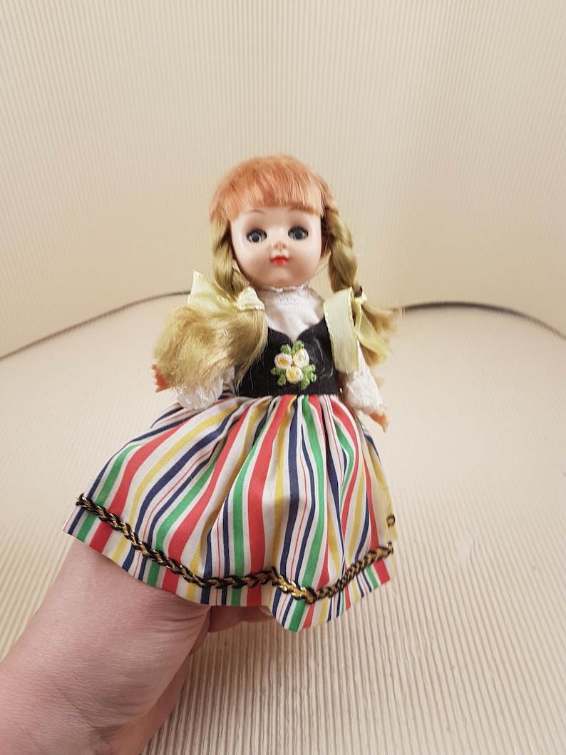 May include: A vintage doll with blonde hair in pigtails, wearing a white blouse, a black bodice with embroidered flowers, and a colorful striped skirt with gold trim.
