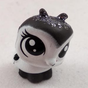 Puede incluir: Pequeña figura pintada a mano de una criatura parecida a un panda. La figura tiene una cara blanca con grandes ojos negros y una cabeza negra con detalles de purpurina. El cuerpo es negro y blanco, con pequeñas orejas.