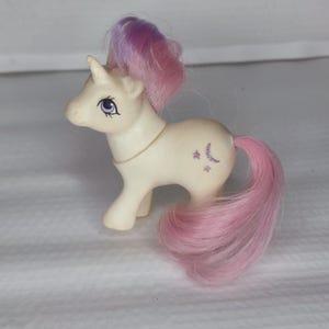 May include: A white unicorn My Little Pony toy with a pink and purple mane and tail. The pony has a crescent moon and stars on its flank.