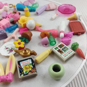 Puede incluir: Una colecci&oacute;n de juguetes en miniatura, que incluye un libro, una zanahoria, un bolo y un peque&ntilde;o jarr&oacute;n verde. Otros art&iacute;culos incluyen un plato con un dise&ntilde;o floral, una flor amarilla y una tetera en miniatura. Los juguetes est&aacute;n dispuestos sobre una superficie blanca.