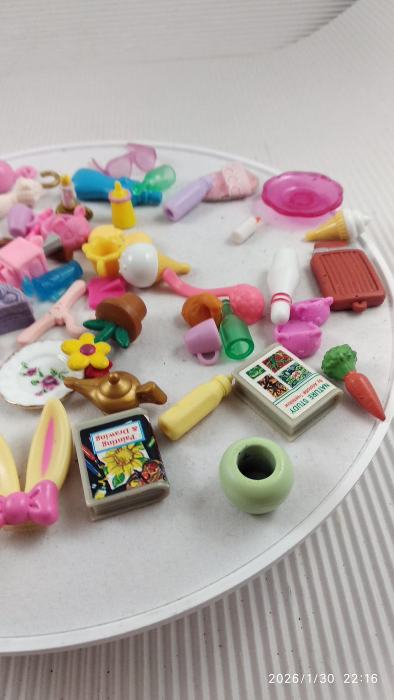 Puede incluir: Una colecci&oacute;n de juguetes en miniatura, que incluye un libro, una zanahoria, un bolo y un peque&ntilde;o jarr&oacute;n verde. Otros art&iacute;culos incluyen un plato con un dise&ntilde;o floral, una flor amarilla y una tetera en miniatura. Los juguetes est&aacute;n dispuestos sobre una superficie blanca.