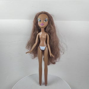 May include: A brown-skinned fashion doll with long brown hair, green eyes, and pink lips. The doll is wearing a white bikini bottom. The doll is standing on a white surface.