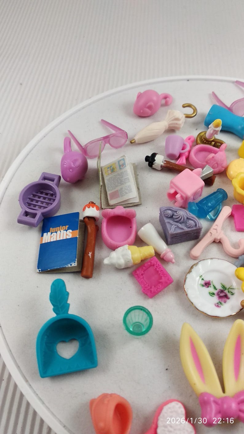 Puede incluir: Colecci&oacute;n de juguetes en miniatura de varios colores, incluyendo rosa, morado, azul y amarillo. Los art&iacute;culos incluyen gafas, un libro con "Junior Maths", una pala y otros peque&ntilde;os accesorios. Los juguetes est&aacute;n dispuestos sobre una superficie blanca.