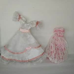 Puede incluir: Dos vestidos de muñeca. Uno es blanco con cinta rosa y ribete de encaje, el otro es rosa con superposición de encaje blanco. Ambos vestidos tienen una estética vintage, con detalles delicados y un aire romántico.