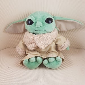 May include: A green plush toy with large eyes and a brown fur coat. The toy is sitting on a white surface.