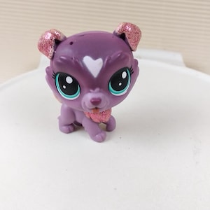 Può includere: Giocattolo per cani di colore viola e rosa glitterato con un cuore bianco sul petto. Il giocattolo ha gli occhi azzurri e una lingua rosa.