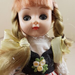May include: A vintage porcelain doll with blonde hair in braids, wearing a black and white dress with a floral design and a colorful striped skirt.