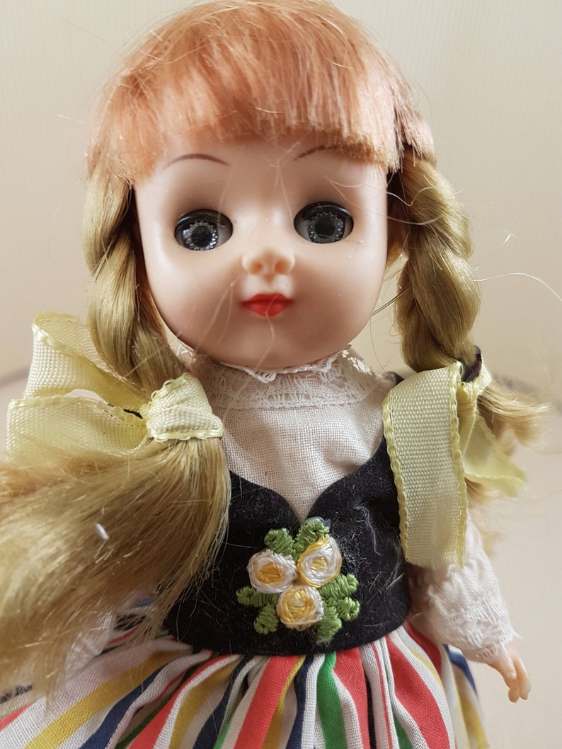 May include: A vintage porcelain doll with blonde hair in braids, wearing a black and white dress with a floral design and a colorful striped skirt.