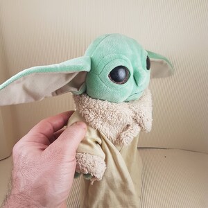 May include: A green plush toy with large eyes and a brown fur coat. The toy is wearing a tan robe.