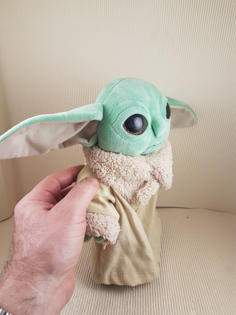 May include: A green plush toy with large eyes and a brown fur coat. The toy is wearing a tan robe.