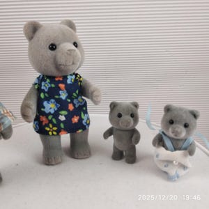 May include: A collection of grey bear figurines. One bear is dressed in a blue floral dress, another has a blue ribbon and diaper. A third bear stands upright. These figurines are likely toys or collectibles.