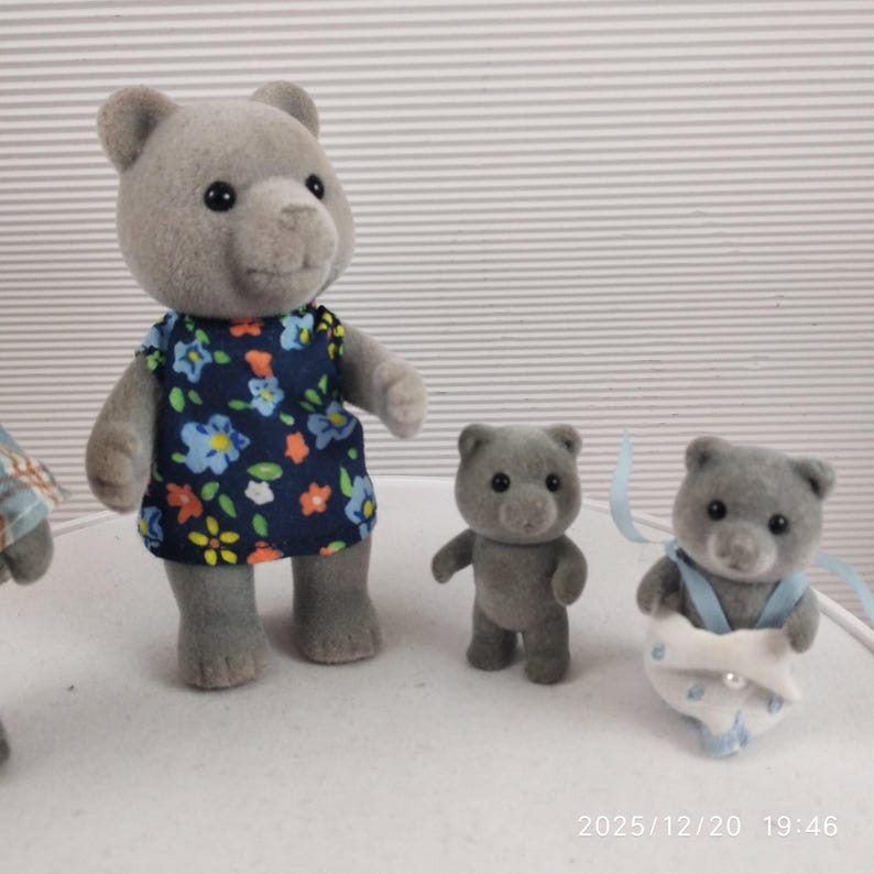 May include: A collection of grey bear figurines. One bear is dressed in a blue floral dress, another has a blue ribbon and diaper. A third bear stands upright. These figurines are likely toys or collectibles.