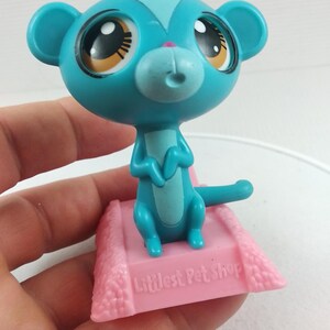 May include: A teal-colored Littlest Pet Shop monkey figurine with large eyes and a pink nose. The monkey is perched on a pink base with the text "Littlest Pet Shop". The figurine is a collectible toy.