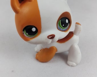 May include: A small, plastic toy cat figurine with a white body and orange markings on its ears, head, and paws. The cat has large, expressive green eyes and a small, upturned tail. The toy is designed in a cartoonish style.