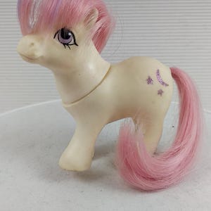 May include: A vintage toy pony figurine in off-white with a pink and purple mane and tail. The pony has purple eyes and glittery moon and star accents on its flank. A classic toy.