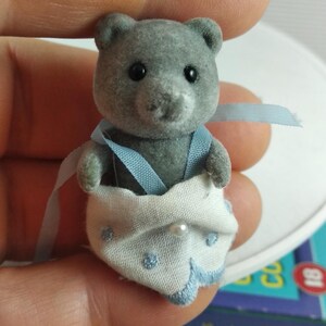 May include: A grey toy bear wearing a white nappy with blue embroidered details and a pearl. The bear has black eyes and is wearing blue braces. The toy is held by a person.