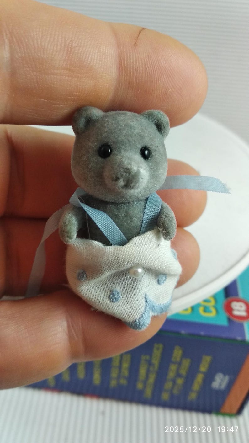 May include: A grey toy bear wearing a white nappy with blue embroidered details and a pearl. The bear has black eyes and is wearing blue braces. The toy is held by a person.