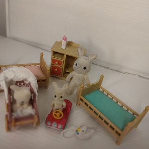 May include: A set of miniature furniture for a dollhouse, including two beds, a dresser, a stroller, a toy car, and two white bunny figurines. The furniture is made of wood and painted in light brown and white.