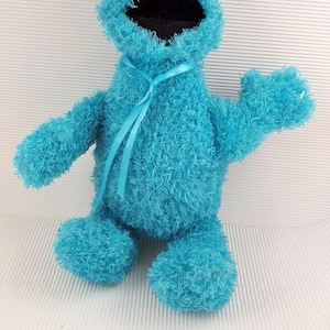 Könnte beinhalten: Ein hellblaues Cookie Monster Plüschtier mit großen weißen Augen und schwarzen Pupillen. Das Spielzeug hat einen weit geöffneten Mund und eine zottelige Textur. Um den Hals ist ein blaues Band gebunden.