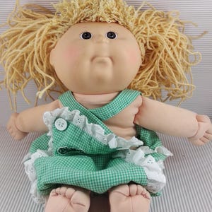 Peut inclure: Une poupée vintage avec un corps en tissu doux et un visage en vinyle. La poupée a des cheveux blonds en fil et porte une robe à carreaux verts et blancs avec une bordure en dentelle blanche. Les bras et les jambes de la poupée sont en tissu doux.