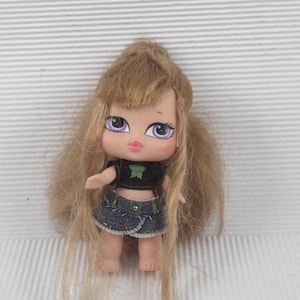 Può includere: Una piccola bambola con lunghi capelli biondi e grandi occhi viola. La bambola indossa un top nero con un disegno verde e una gonna di jeans. La bambola è in piedi con le braccia lungo i fianchi.