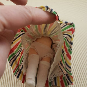May include: A doll wearing a white dress with a colorful striped skirt and white socks.
