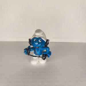 May include: A blue Smurf figurine wearing white headphones and holding a black cassette player. The Smurf is smiling and has a white hat.