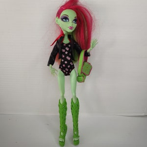 Peut inclure: Une poupée Monster High verte avec des cheveux roses et verts portant un maillot de bain noir et blanc, une veste en cuir noir et des sandales de gladiateur vertes. Elle tient un sac à main vert et rose.