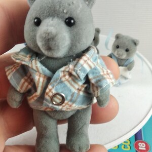 May include: A grey teddy bear figurine wearing a blue and brown plaid shirt. Other bear figurines are in the background. The bear is a toy or collectible item. The image also shows a DVD case with the number 18.