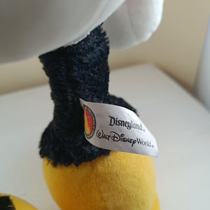 May include: A white plush toy with black legs and yellow feet. The toy has a white tag attached to one leg that reads "Disneyland. Walt Disney World."
