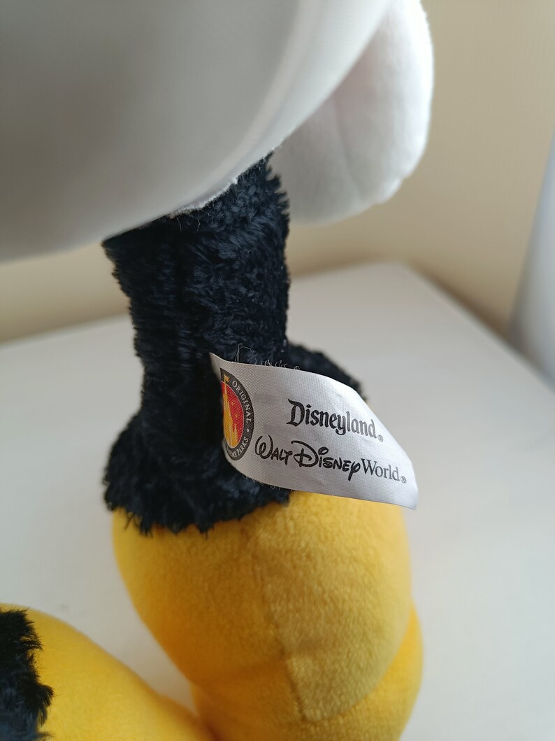 May include: A white plush toy with black legs and yellow feet. The toy has a white tag attached to one leg that reads "Disneyland. Walt Disney World."