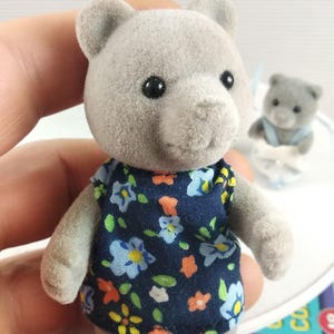 May include: A grey teddy bear figure wearing a navy blue dress with a floral pattern of blue, yellow, and orange flowers. Another bear is visible in the background. The bear is held by a person.