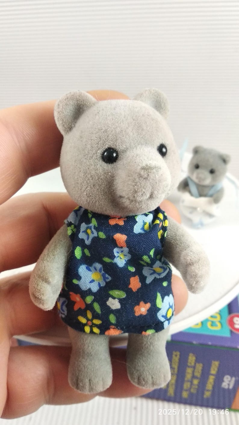 May include: A grey teddy bear figure wearing a navy blue dress with a floral pattern of blue, yellow, and orange flowers. Another bear is visible in the background. The bear is held by a person.