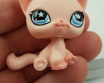 May include: A light pink cat figurine with large blue eyes and star-shaped pupils. The cat has small pink ears, a tiny pink nose, and a curled tail. The figurine is held in a hand, showcasing its small size and detailed features.