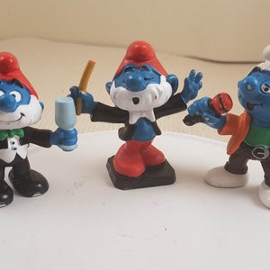 May include: Three Smurf figurines in different poses. One wears a tuxedo and holds a glass, another conducts with a baton, and the third holds a microphone and points. Each figure has a blue body, white beard, and a red or white hat.