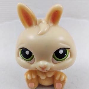 May include: A cream-coloured bunny figurine with large green eyes and pink accents on the ears and paws. The toy bunny has a sweet expression and is posed in a sitting position. The figurine is a collectible toy.