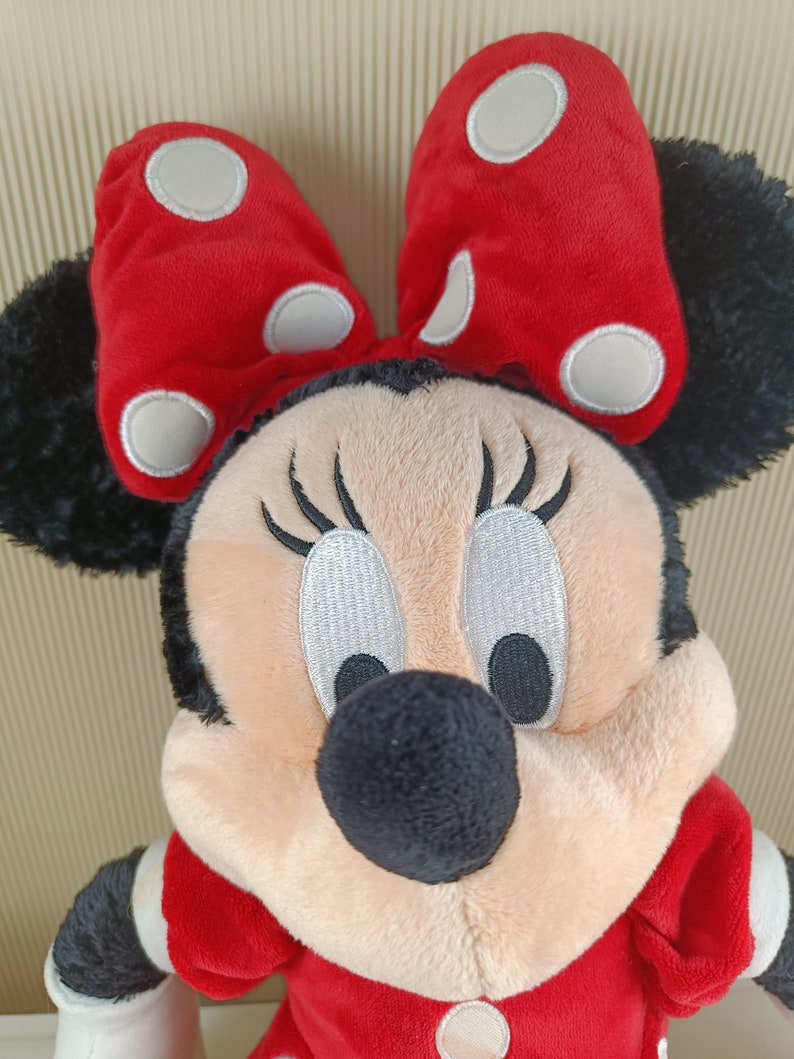 May include: A plush toy of Minnie Mouse wearing a red dress with white polka dots and a red bow with white polka dots. She has black ears, a black nose, and black eyes with white eyelashes.